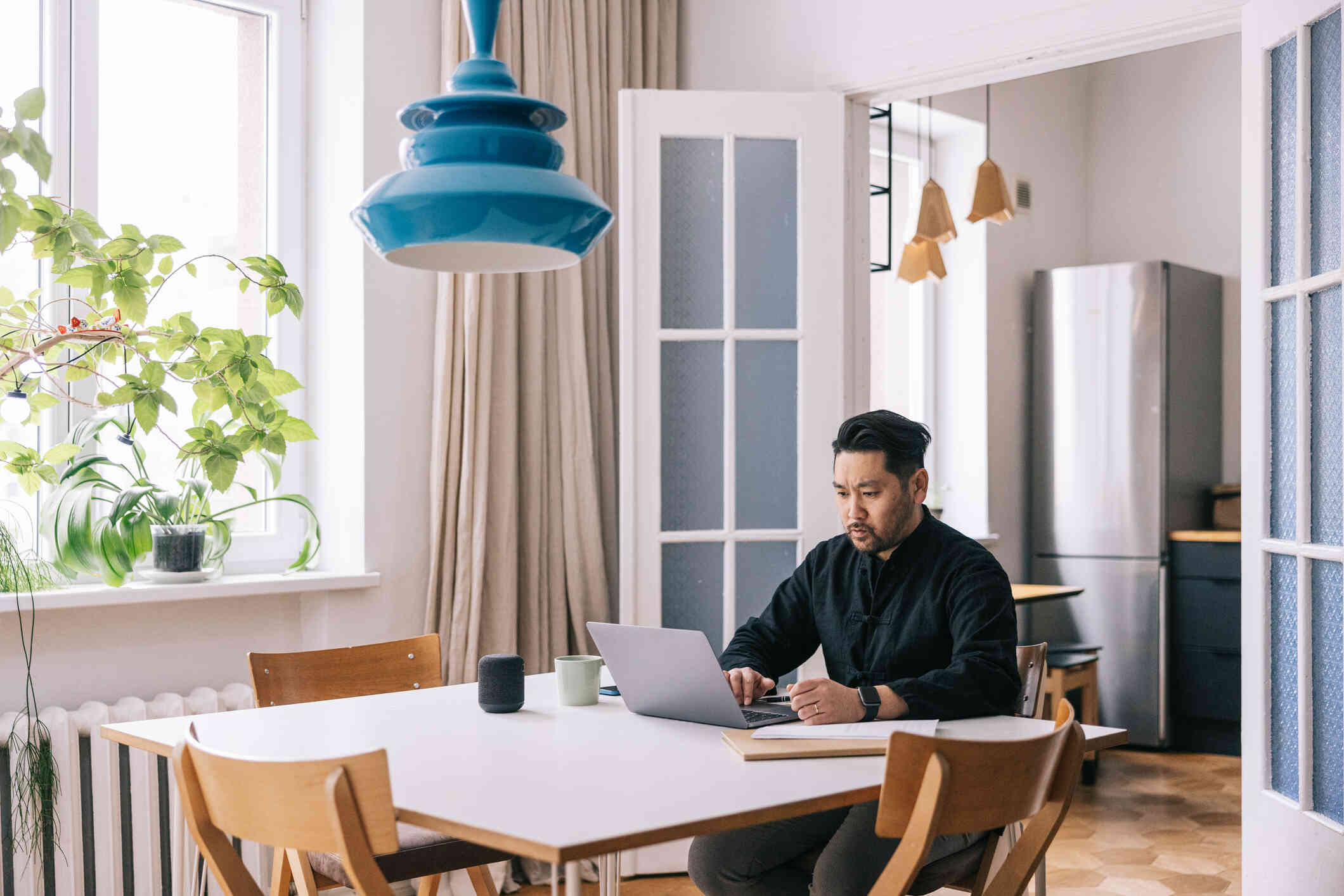 A man in a black shirt sits at his kitchen table and looks at the laptop open infront of him with a serious expression.