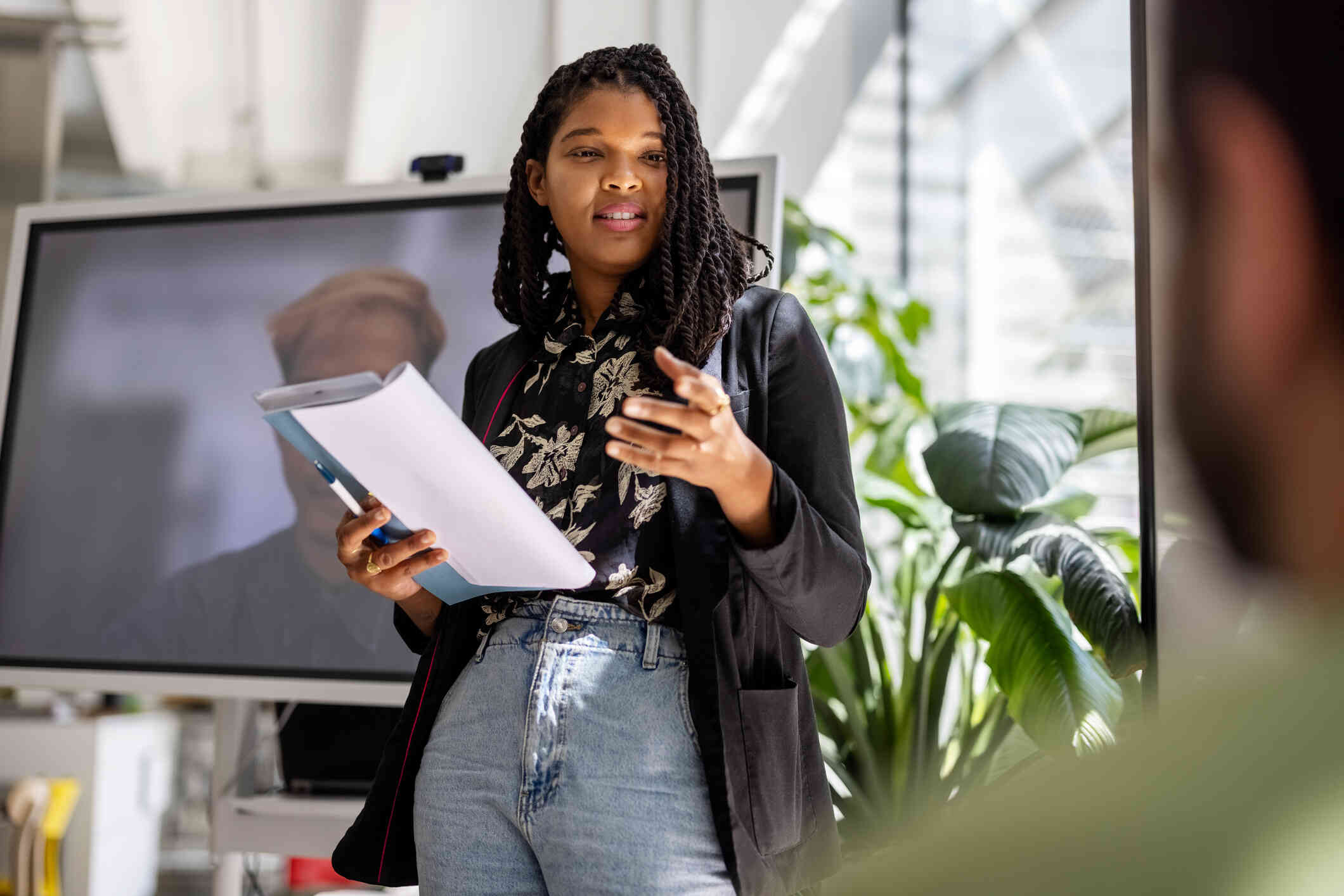 A woman ina black top stands at the front of a room and elads a meeting while holding a book in her hands.