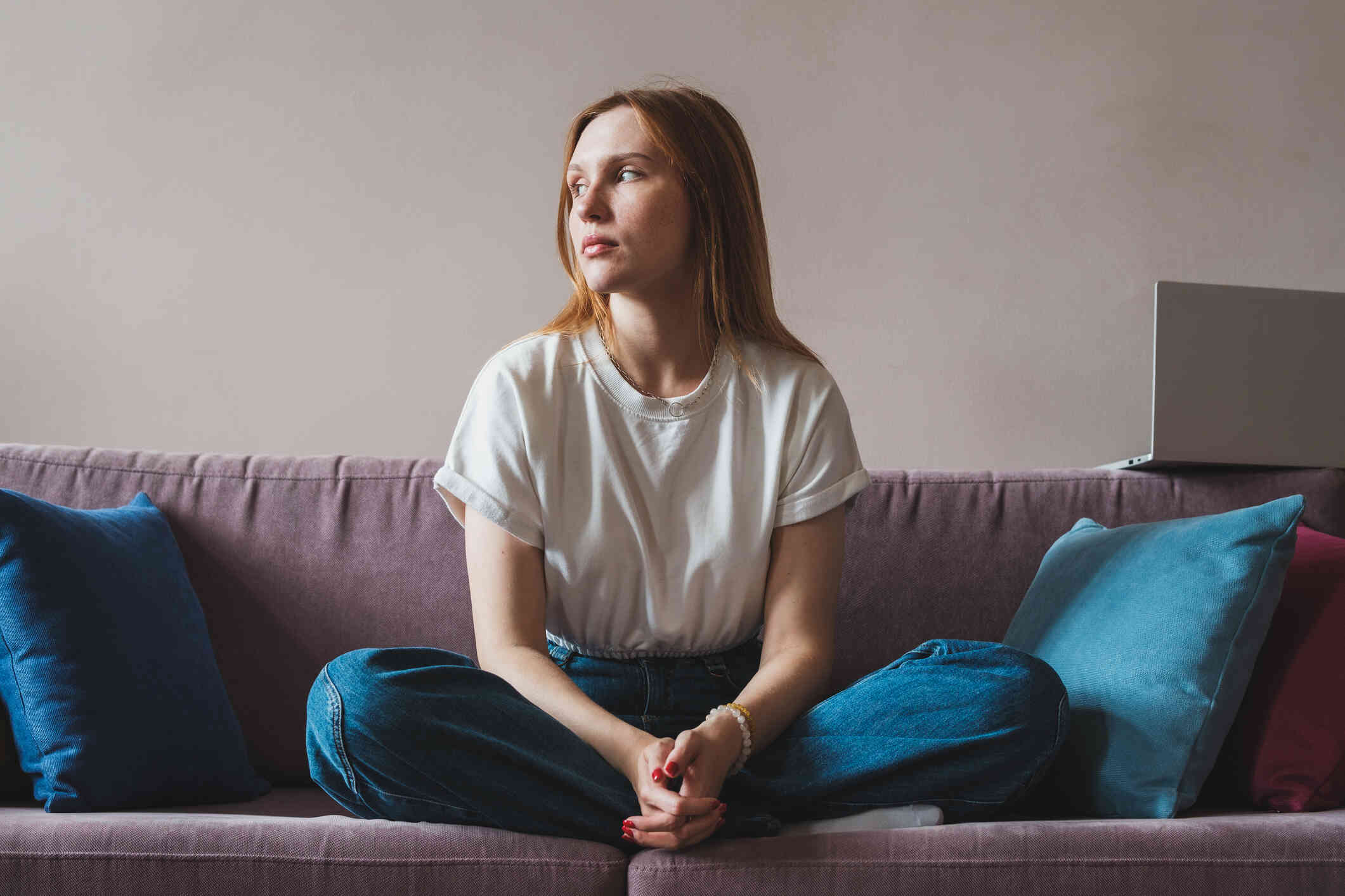 A woman in a white shirt sits cross legged on her couch while gazing off sadly.