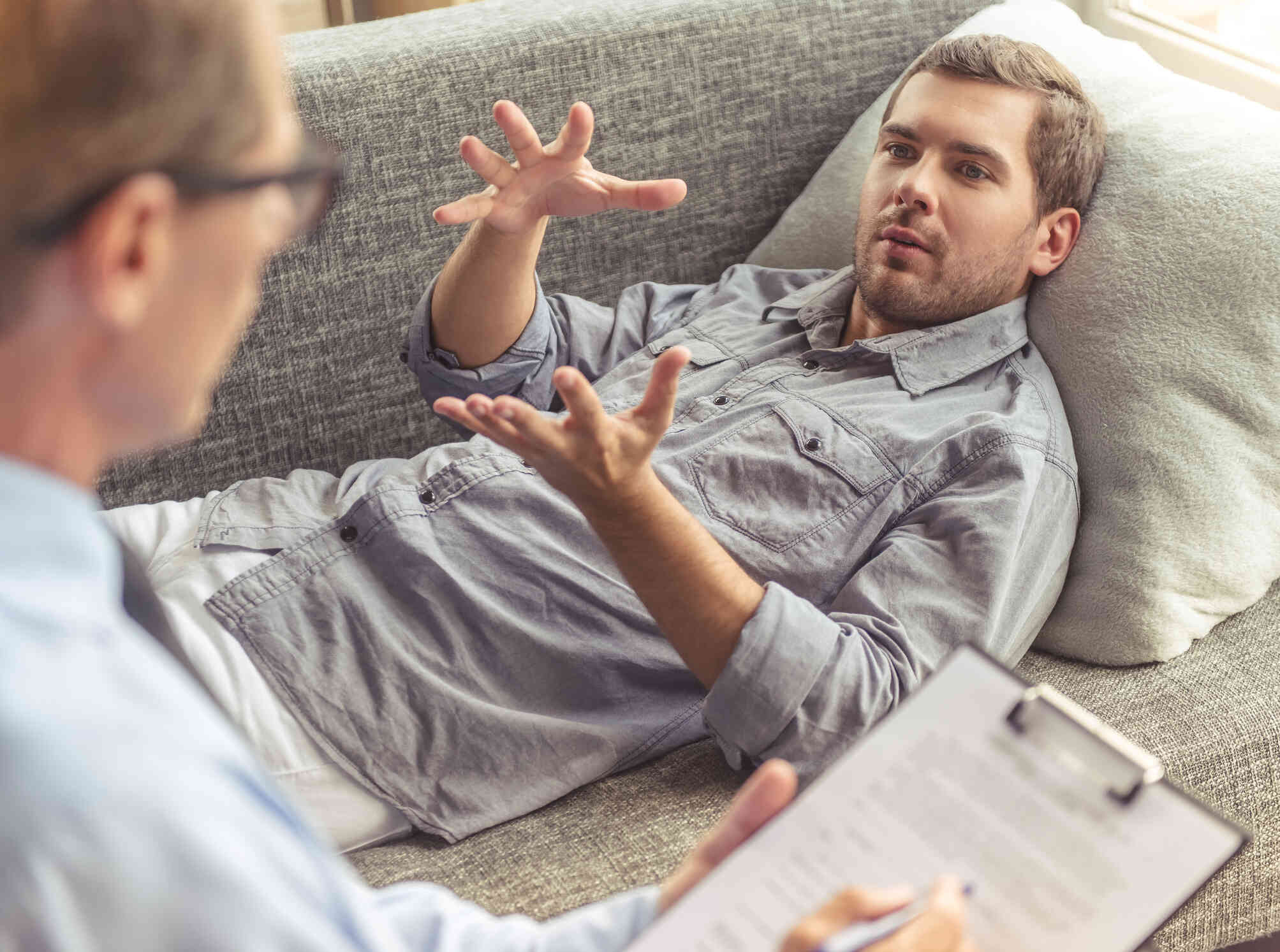 A man in a grey shirt lays on his back on a couch while talking to the male therapist sitting next to him during a therapy session.