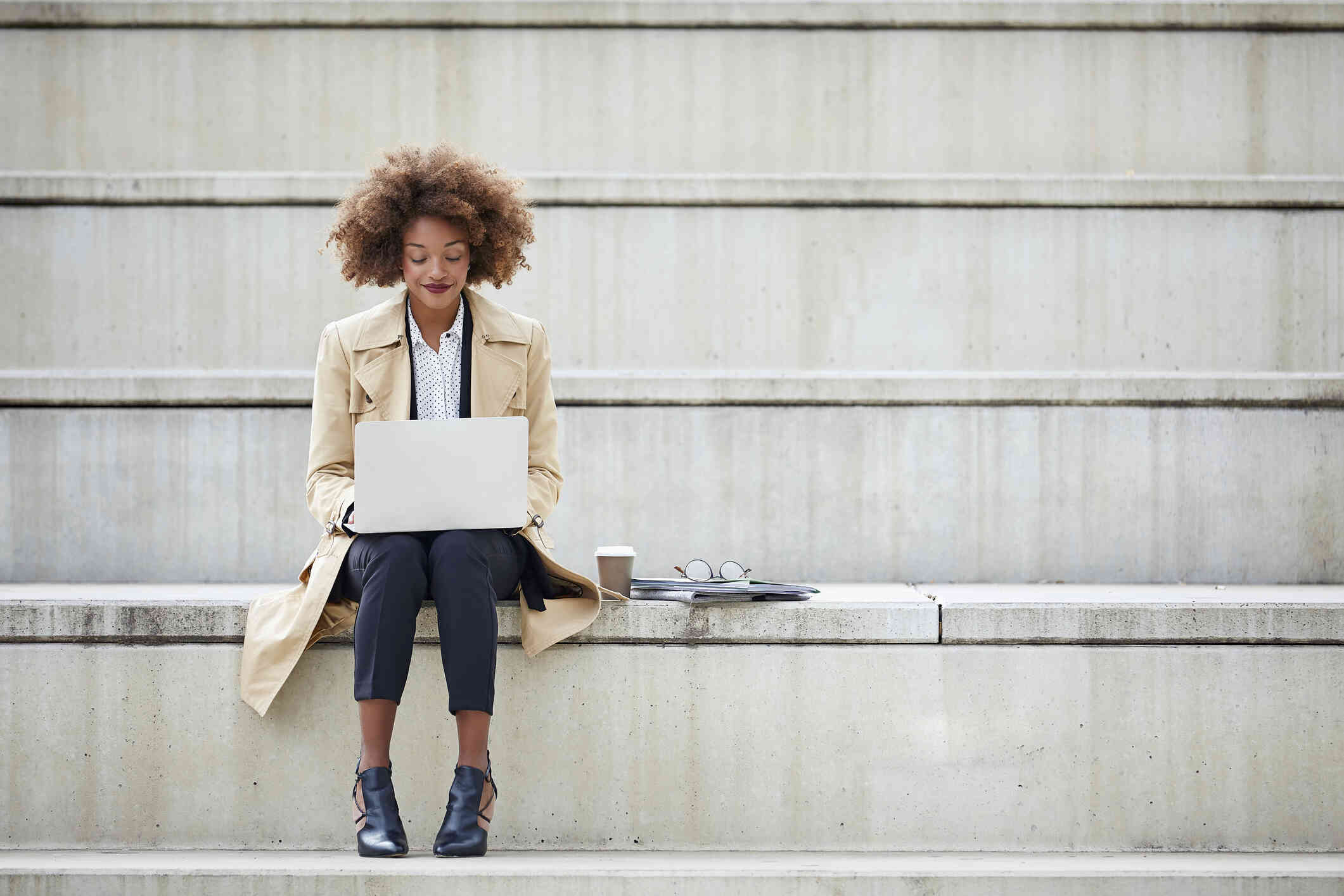 A woman in a tan coat sits on a cement step with her laptop in her lap as she types on the keyboard.